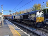 Freight train under electric overhead wires passes a passenger station.