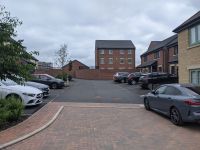 Cars parked around a large piece of land with tarmac and paved covering. Houses surround the parking area.