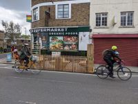 In front of a convenience store are two cyclists passing a parklet with seating.