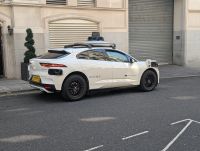 On a city street, a white car with sensors and other equipment attached to it. Some branding reads "Waymo".